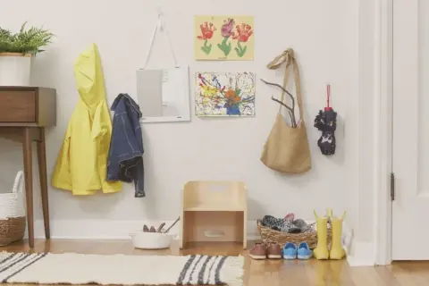 Montessori entryway ideas: a yellow raincoat, denim jacket, and artwork hang on the wall. A small wooden stool and baskets hold shoes and boots.
