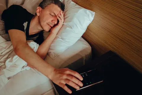 Man in bed, hand reaching for phone alarm. Does snoozing make you more tired?