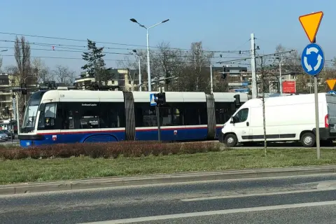 Tramwaj na rondzie, obok biała furgonetka. Czy tramwaj ma pierwszeństwo na rondzie? Znak informuje o ruchu okrężnym.