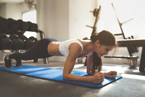 Woman performing a plank jack exercise on a blue mat in a gym, with weights in the background.