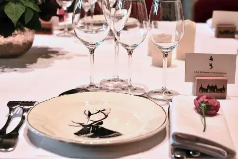 Une table dressée pour un dîner de gala, avec une assiette ornée d'un cerf, des verres à vin et une carte de visite "S.A.R. LE Grand-Duc de Luxembourg". La fourchette à la française est prête pour le festin.