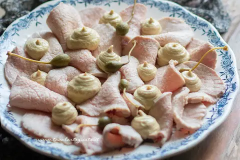 Vitello tonnato della nonna: fette sottili di carne rosa, guarnite con salsa tonnata e capperi, su un piatto bianco con decori blu.