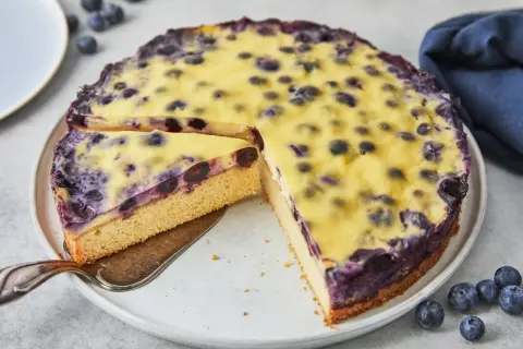 Ein saftiger Heidelbeerkuchen mit cremigem Vanillepudding, ein Stück wird gerade herausgehoben.