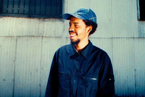 Earl Sweatshirt, smiling and wearing a blue plaid cap and dark jacket, stands against a weathered wall. He's ready to drop his next earl sweatshirt setlist.