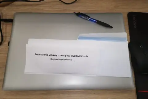 Na laptopie leżą dokumenty z napisem "Rozwiązanie umowy o pracę bez wypowiedzenia (Zwolnienie dyscyplinarne)". Obok długopis i myszka.