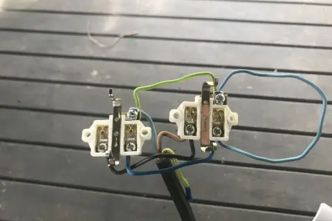 Mostkowanie podwójnego gniazdka elektrycznego. Widać białe elementy z zaciskami i kolorowe przewody: zielony, niebieski i brązowy.