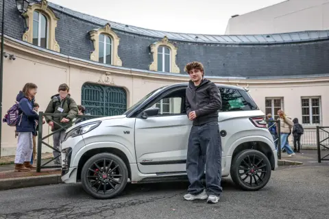 Un jeune homme pose à côté d'une voiturette électrique blanche. Le **rayon de braquage** de ce véhicule compact est idéal pour les rues étroites.