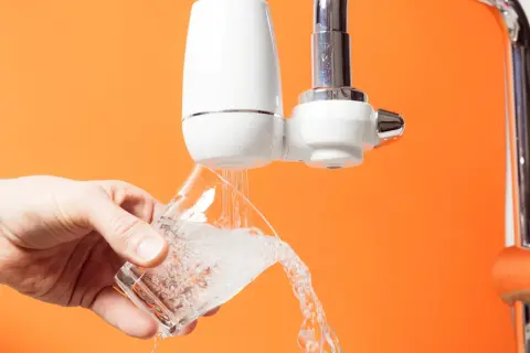 Un robinet avec un filtre blanc verse de l'eau dans un verre. C'est le moment de changer le filtre robinet pour une eau plus pure.