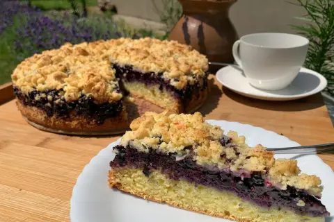 Ein saftiger Heidelbeerkuchen mit Streuseln, ein Stück auf einem Teller, daneben eine Tasse Tee.