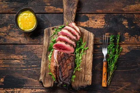 Perfekt gegarte Entenbrust mit rosa Kern, auf einem Holzbrett mit Rucola, dazu eine Schale mit Sauce und ein Gabel. Die Kerntemperatur der Entenbrust ist ideal.