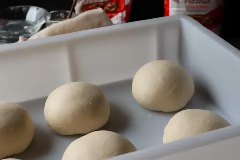 Mehrere Teigkugeln liegen in einer weißen Box, bereit für das Pizzateig einfrieren. Im Hintergrund sind Zutaten und eine Pizzatüte zu sehen.