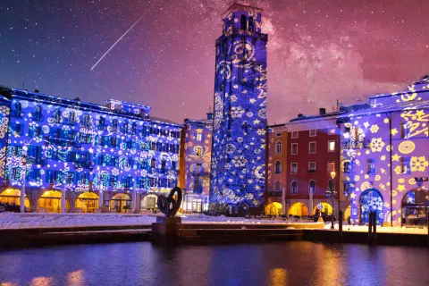 Edifici illuminati da proiezioni natalizie sul lago di Garda a dicembre, con cielo stellato e una scia luminosa.