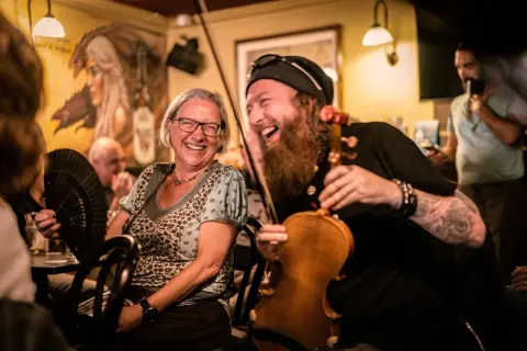 Una mujer ríe mientras un violinista con barba toca. El ambiente es festivo, ¡qué buen craic!