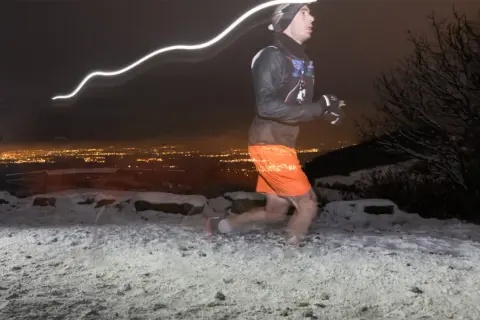 Un coureur nocturne s'entraîne pour la Saintélyon sur un sentier enneigé, la ville illuminée en arrière-plan.