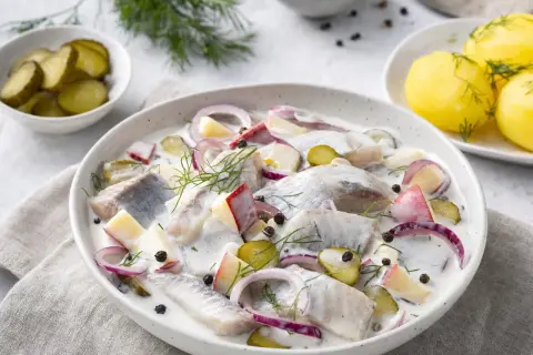 Marinierter Hering mit Äpfeln, Zwiebeln und Dill in cremiger Sauce, garniert mit Pfefferkörnern. Dazu Pellkartoffeln und Gewürzgurken.