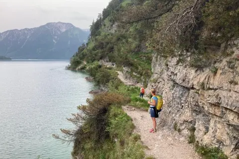 Wanderer genießen die Achensee Umrundung auf einem schmalen Pfad entlang steiler Felswände mit Blick auf den See und die Berge.