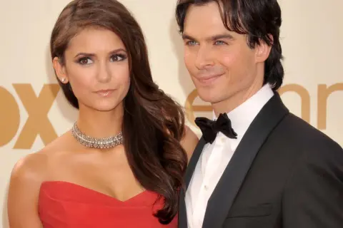Nina Dobrev w czerwonej sukni i jej partner w smokingu, uśmiechnięci na ściance.
