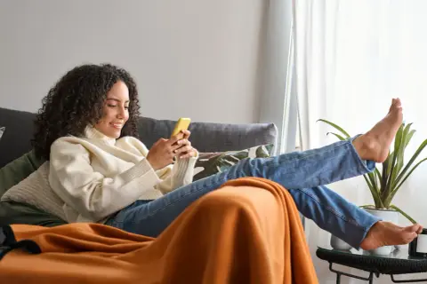Frau mit lockigen Haaren entspannt sich richtig sitzend auf dem Sofa, schaut auf ihr Handy.