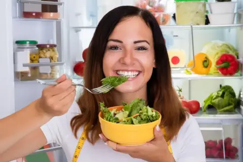 Uśmiechnięta kobieta je sałatkę z lodówki, wiedząc, że zdrowe jedzenie to klucz, jak przyspieszyć metabolizm.
