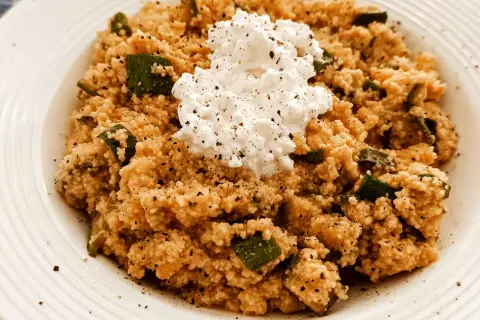 Un piatto di cous cous con zucchine e una generosa porzione di fiocchi di latte al centro, condito con pepe nero.