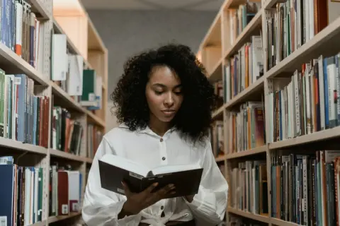 Młoda kobieta z burzą loków czyta książkę w bibliotece. Szuka inspiracji, co napisać we wniosku o stypendium szkolne.