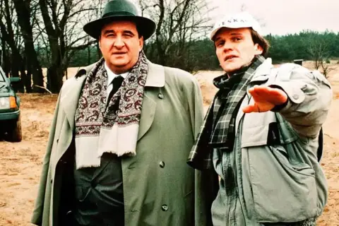 Dwóch mężczyzn w płaszczach i szalikach, jeden w kapeluszu, drugi w czapce. W tle drzewa i piasek. Nic śmiesznego.