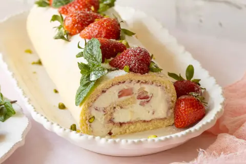 Eine köstliche Erdbeer Biskuitrolle, verziert mit frischen Erdbeeren, Minze und Pistazien, auf einem weißen Teller.