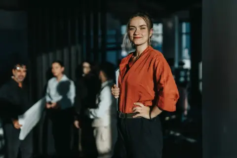 Uśmiechnięta kobieta w pomarańczowej bluzce trzyma teczkę, jakby właśnie odebrała prawo jazdy. W tle grupa osób.