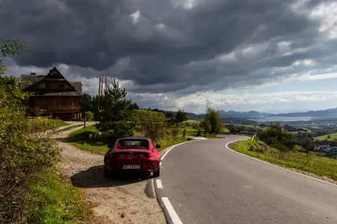Czerwony kabriolet zaparkowany przy drewnianym domu. Droga prowadzi przez malownicze wzgórza, idealne miejsca, co zwiedzić po drodze do Zakopanego.