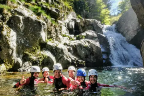 Groupe souriant en combinaison néoprène, casques blancs et bleus, profitant d'une descente en canyoning près d'une cascade. Une belle aventure de canyoning dans le Vallon Pont d'Arc.