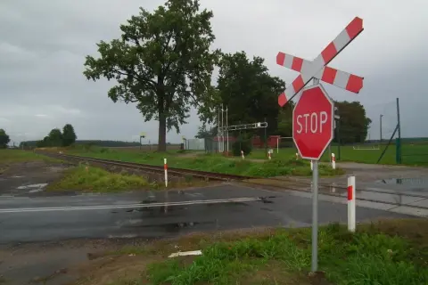 Przejazd kolejowy z zaporami i znakiem STOP. Torowisko przecina mokrą drogę, a w tle widać drzewa i boisko.