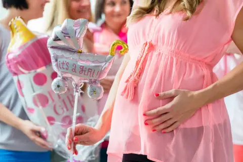 Ciężarna kobieta w różowej sukience trzyma balon w kształcie wózka z napisem "it's a girl!".