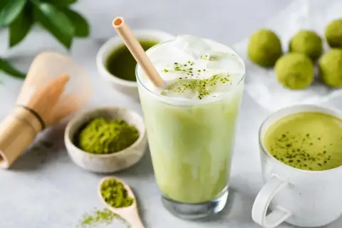 Orzeźwiający iced matcha latte z pianką i bambusową słomką, obok miseczki z matchą i tradycyjnej miotełki do jej przygotowania.