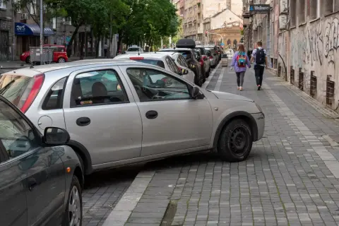 Samochody na chodniku utrudniają przejście. Dwie osoby idą wąskim chodnikiem, omijając przeszkody.