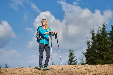 Kobieta z plecakiem i kijkami trekkingowymi na górskim szlaku. Zastanawia się, jak przymocować kijki do plecaka, by mieć wolne ręce.