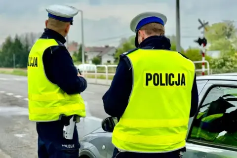 Dwóch policjantów w kamizelkach odblaskowych przy samochodzie. Jeden z nich trzyma broń. Prawdopodobnie zatrzymali kierowcę za przekroczenie prędkości o 50 km/h.