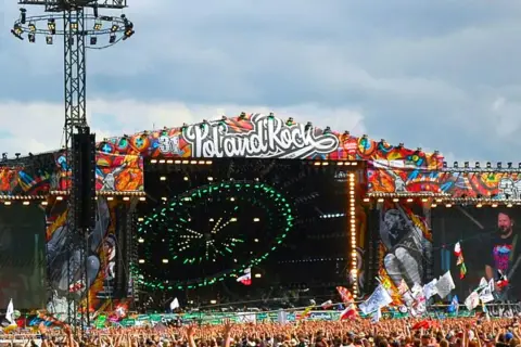 Ogromna scena festiwalowa z napisem "Poland Rock" i kolorowymi grafikami. Tłum ludzi czeka na koncert.