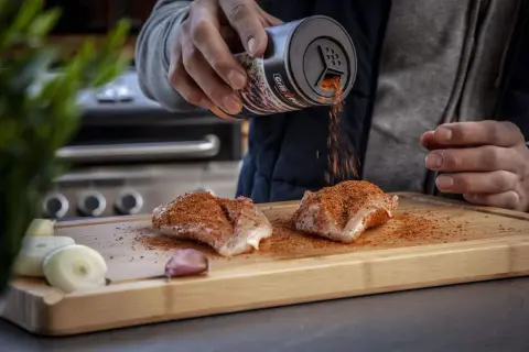 Ein Mann bestreut Hähnchenbrust mit einem magischen Gewürz-Rub auf einem Holzbrett. Zwiebeln und Knoblauch liegen daneben.