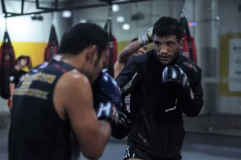 Two men in boxing gloves engage in a spar fight in a gym, with punching bags in the background.