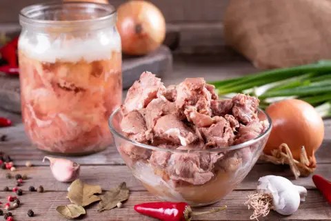 Słoik z domowym mięsem, jak u babci, pełen smaku. Obok miska z porcjami mięsa, cebula, czosnek, papryczki chili i przyprawy.