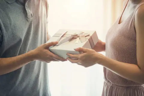 Mężczyzna wręcza kobiecie prezent z okazji 25 rocznicy ślubu. Delikatne wstążki zdobią pudełko.