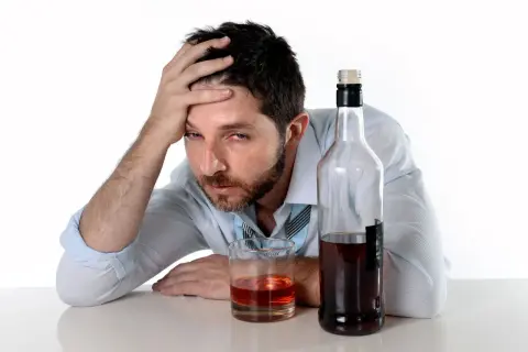 Mężczyzna z ręką na czole, obok szklanka i butelka alkoholu, sugerujące zatrucie alkoholowe.
