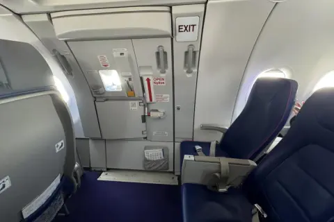 Wnętrze samolotu Wizzair. Widok na rząd siedzeń i drzwi wyjściowe. Numeracja miejsc w samolocie Wizzair jest widoczna na siedzeniach.
