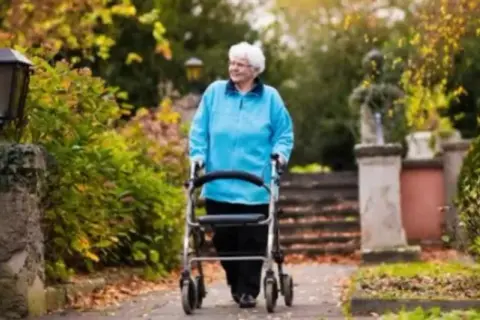 Elderly woman in blue jacket walks with a walker in a park with autumn foliage.