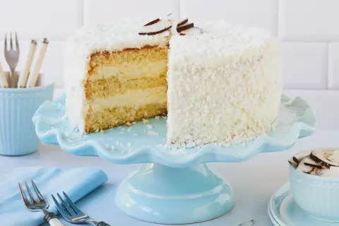 Eine köstliche Kokos Torte, bedeckt mit Kokosraspeln und dekoriert mit Kokoschips, wartet auf dem hellblauen Tortenständer.