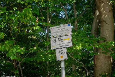 Znaki szlaku rowerowego wskazują trasę 003 do Starego Bielska, 17 km. W tle zielone drzewa.