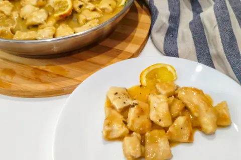 Petto di pollo al limone cremoso servito su un piatto bianco, con una fetta di limone e una padella piena dello stesso piatto sullo sfondo.