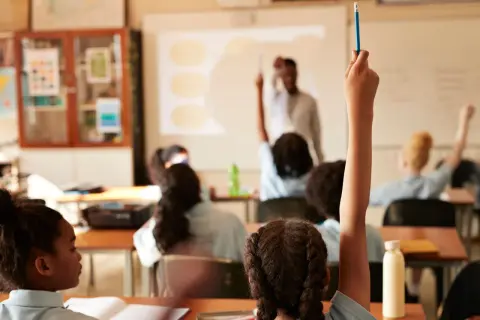 Estudiantes levantan la mano en clase. El profesor proyecta información, quizás sobre el calendario escolar 2026-27.