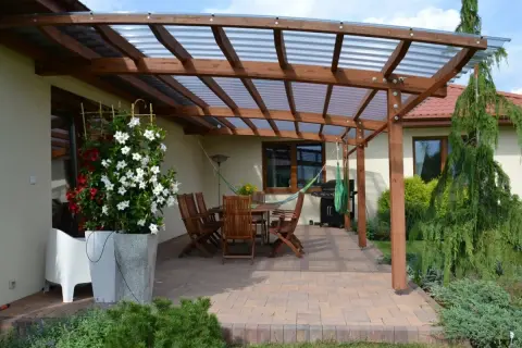 Drewniana pergola z przezroczystym dachem nad tarasem, gdzie stoi stół z krzesłami, grill i hamak. Idealne miejsce, co na taras.