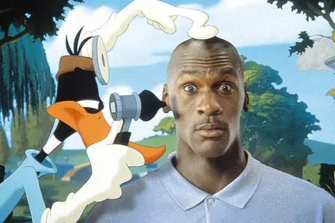 Michael Jordan z zaskoczoną miną, gdy Kaczor Daffy próbuje mu coś włożyć do ucha. Zabawna scena z komedii dla dzieci.